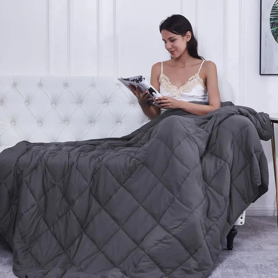 Woman reading a book under a gray quilt on a bed.